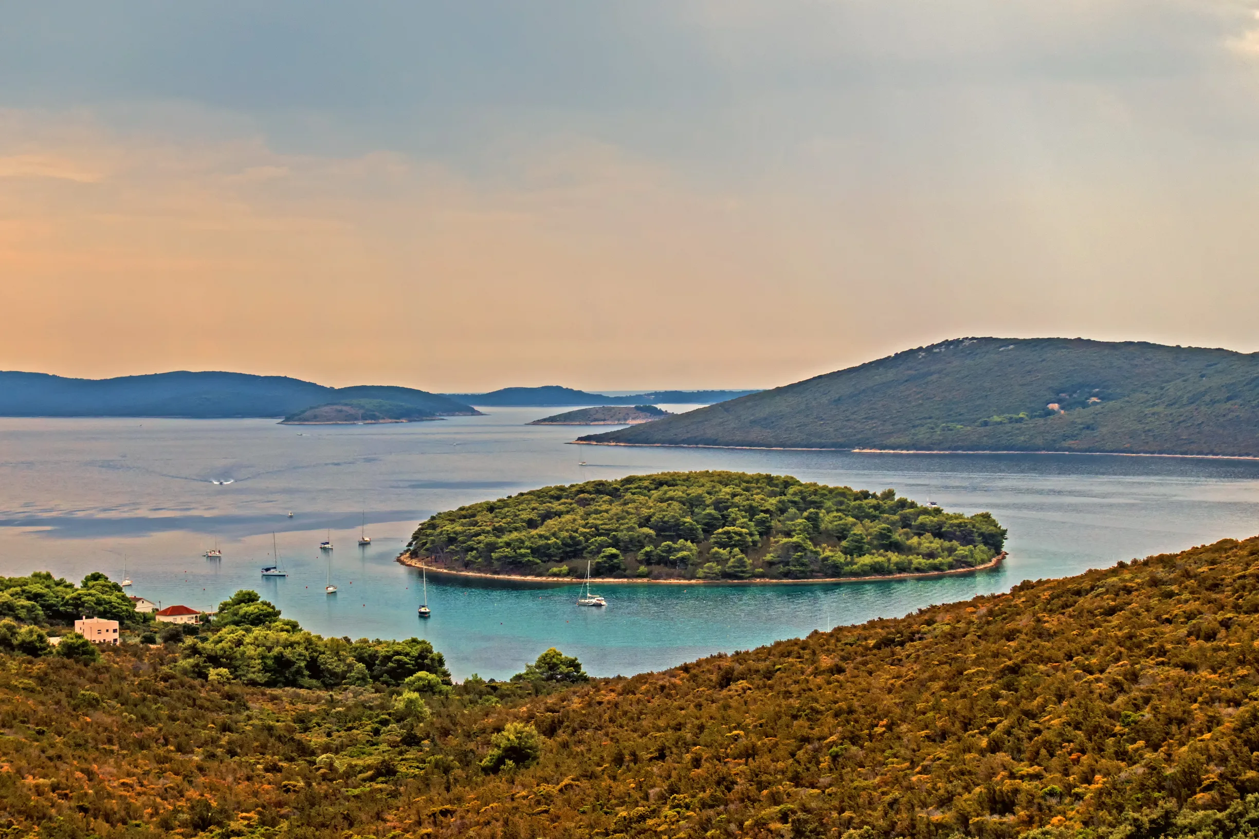 Sailboats anchored around round wooded islet under moody evening sky