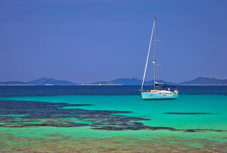 Sailboat anchored in turquoise shallows with low islands in the background